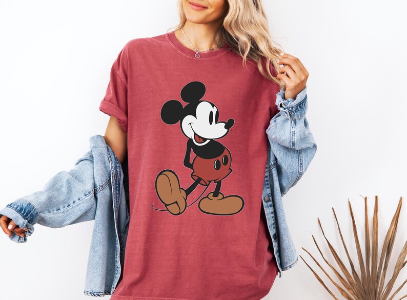 May include: Red t-shirt featuring a classic cartoon Mickey Mouse design. The graphic shows Mickey in his iconic pose, with black ears, a white face, red shorts, and brown shoes. The shirt is a solid red color.