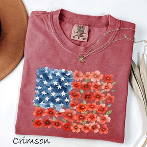 May include: Crimson-coloured t-shirt with a floral American flag design. The flag design features blue watercolour stars and red flowers. The shirt has a crew neck and a small tag near the neckline. A gold necklace is draped across the shirt.