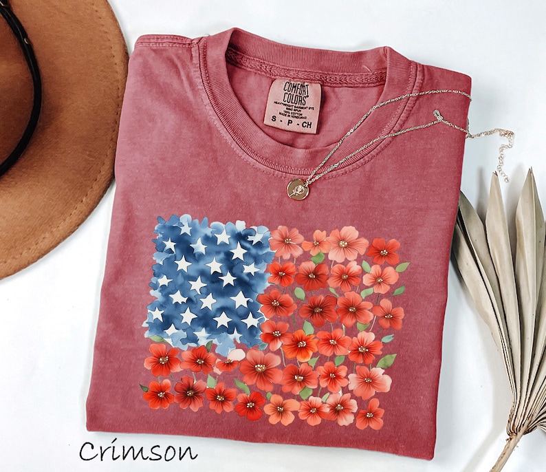May include: Crimson-coloured t-shirt with a floral American flag design. The flag design features blue watercolour stars and red flowers. The shirt has a crew neck and a small tag near the neckline. A gold necklace is draped across the shirt.