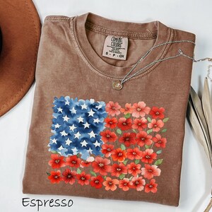 May include: Espresso-coloured t-shirt with a floral American flag design. The flag design features blue watercolour stars and red flowers. The shirt has a crew neck and short sleeves. A silver necklace with a pendant is draped on the shirt.