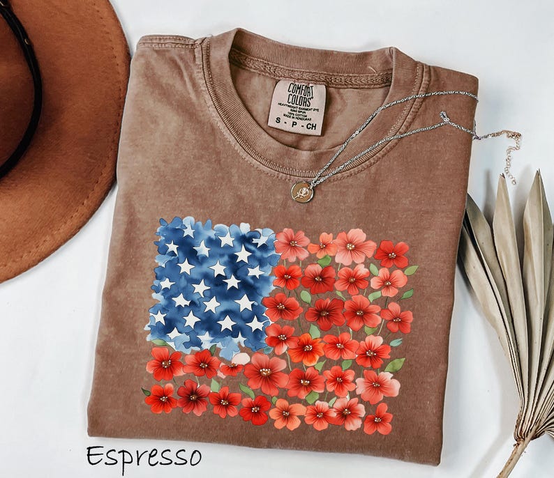May include: Espresso-coloured t-shirt with a floral American flag design. The flag design features blue watercolour stars and red flowers. The shirt has a crew neck and short sleeves. A silver necklace with a pendant is draped on the shirt.