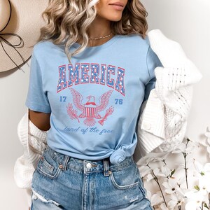 May include: A light blue t-shirt with a red, white, and blue graphic of an eagle and the words "AMERICA 17 76 land of the free".