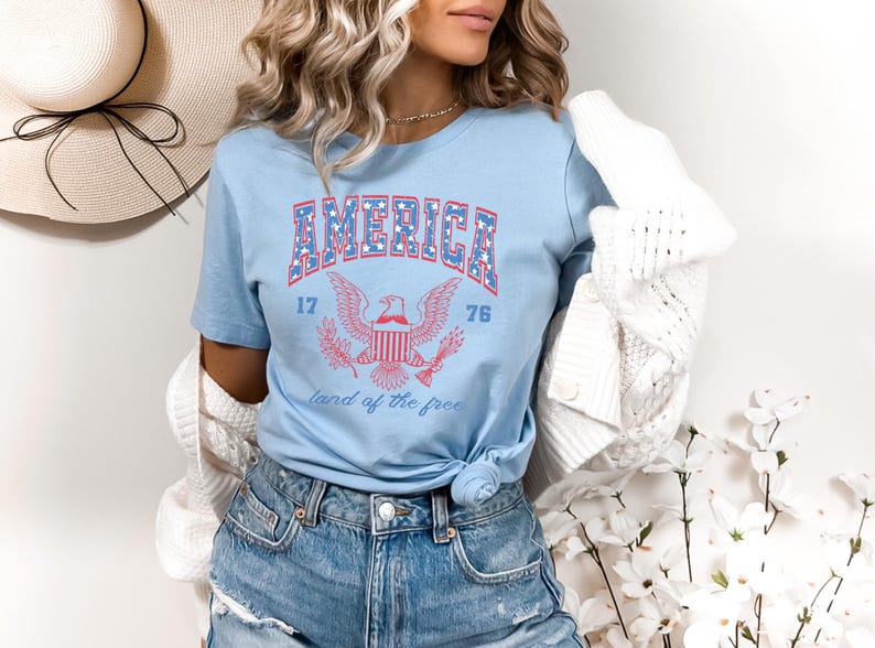 May include: A light blue t-shirt with a red, white, and blue graphic of an eagle and the words "AMERICA 17 76 land of the free".