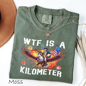 K&ouml;nnte beinhalten: Ein moosgr&uuml;nes T-Shirt mit dem wei&szlig;en Schriftzug "WTF IS A KILOMETER". Die Grafik zeigt einen Soldaten, der auf einem Adler reitet und ein Gewehr und eine amerikanische Flagge h&auml;lt. Eine goldene Halskette liegt &uuml;ber dem Shirt.