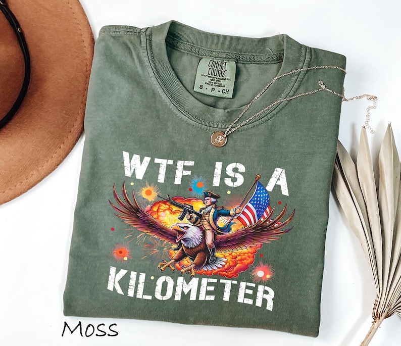 K&ouml;nnte beinhalten: Ein moosgr&uuml;nes T-Shirt mit dem wei&szlig;en Schriftzug "WTF IS A KILOMETER". Die Grafik zeigt einen Soldaten, der auf einem Adler reitet und ein Gewehr und eine amerikanische Flagge h&auml;lt. Eine goldene Halskette liegt &uuml;ber dem Shirt.