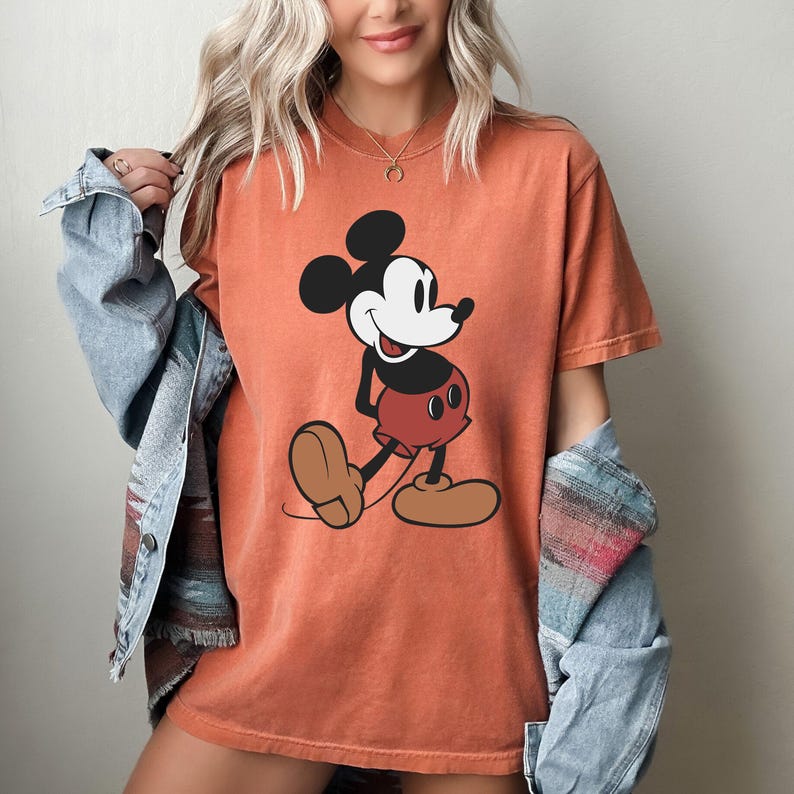 May include: Orange t-shirt featuring a classic Mickey Mouse graphic. The cartoon character is depicted in black, white, red, and brown. The shirt has short sleeves and a crew neck.