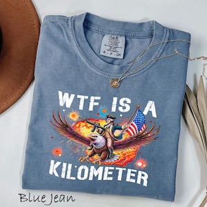 K&ouml;nnte beinhalten: Blaues Jeans-T-Shirt mit dem Schriftzug "WTF IS A KILOMETER" in wei&szlig;en Blockbuchstaben. Die Grafik zeigt einen Mann, der auf einem Adler reitet und ein Gewehr und eine amerikanische Flagge h&auml;lt. Eine goldene Halskette liegt auf dem T-Shirt.