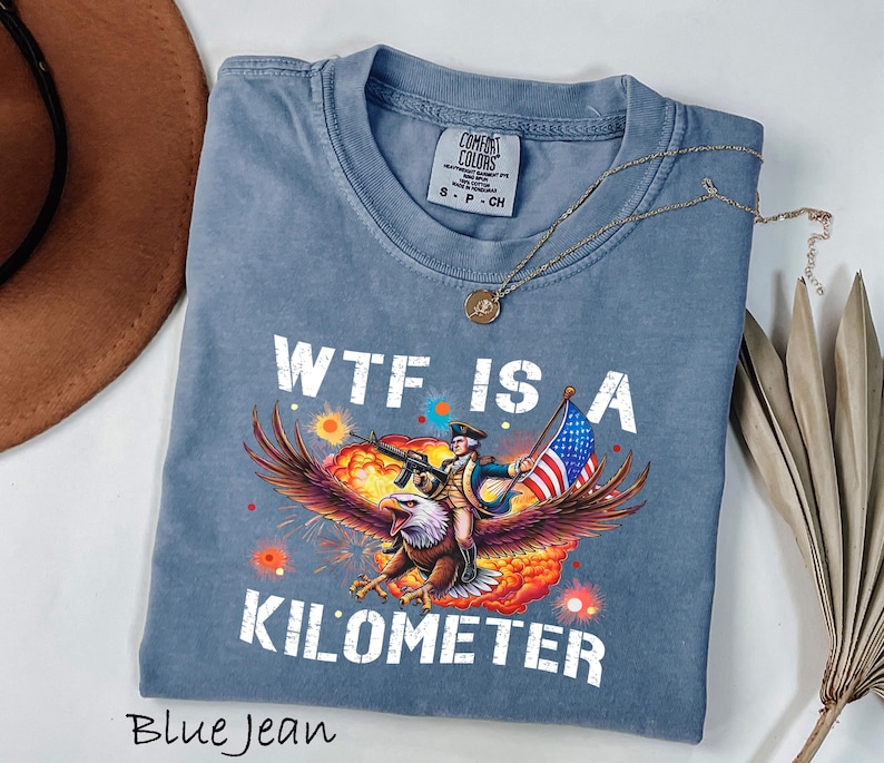 K&ouml;nnte beinhalten: Blaues Jeans-T-Shirt mit dem Schriftzug "WTF IS A KILOMETER" in wei&szlig;en Blockbuchstaben. Die Grafik zeigt einen Mann, der auf einem Adler reitet und ein Gewehr und eine amerikanische Flagge h&auml;lt. Eine goldene Halskette liegt auf dem T-Shirt.