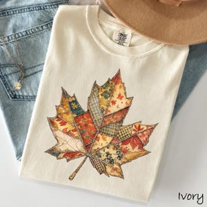 May include: An ivory t-shirt featuring a large maple leaf graphic composed of various patchwork patterns in shades of orange, yellow, green, and brown. The inner neck label reads Comfort Colors. The shirt is displayed with blue denim and a tan hat.