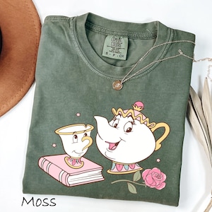 May include: Moss green t-shirt featuring a cartoon illustration of a teacup, teapot, book, and rose. The design is in white, pink, and yellow. A gold necklace with a pendant is on the shirt. The word "Moss" is printed at the bottom.