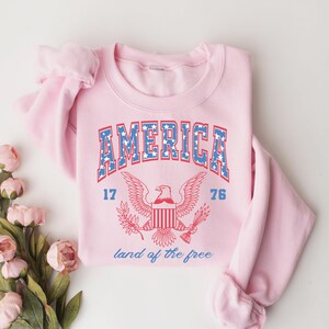 May include: Pink sweatshirt with a vintage-style graphic of an eagle and the text "AMERICA 17 76 land of the free".