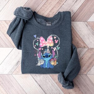 May include: A dark grey sweatshirt with a colourful graphic of the character Stitch from Lilo & Stitch. The design includes a pink bow, castle, and other elements. The sweatshirt is folded on a wooden surface.