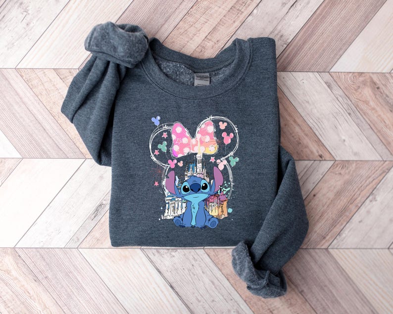 May include: A dark grey sweatshirt with a colourful graphic of the character Stitch from Lilo & Stitch. The design includes a pink bow, castle, and other elements. The sweatshirt is folded on a wooden surface.