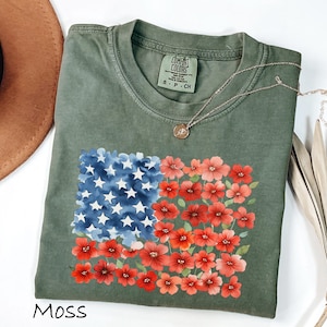 May include: Moss green t-shirt with a floral American flag design. The flag design features blue watercolor stars and red floral stripes. The shirt has a crew neck and short sleeves. A gold necklace is draped across the shirt.