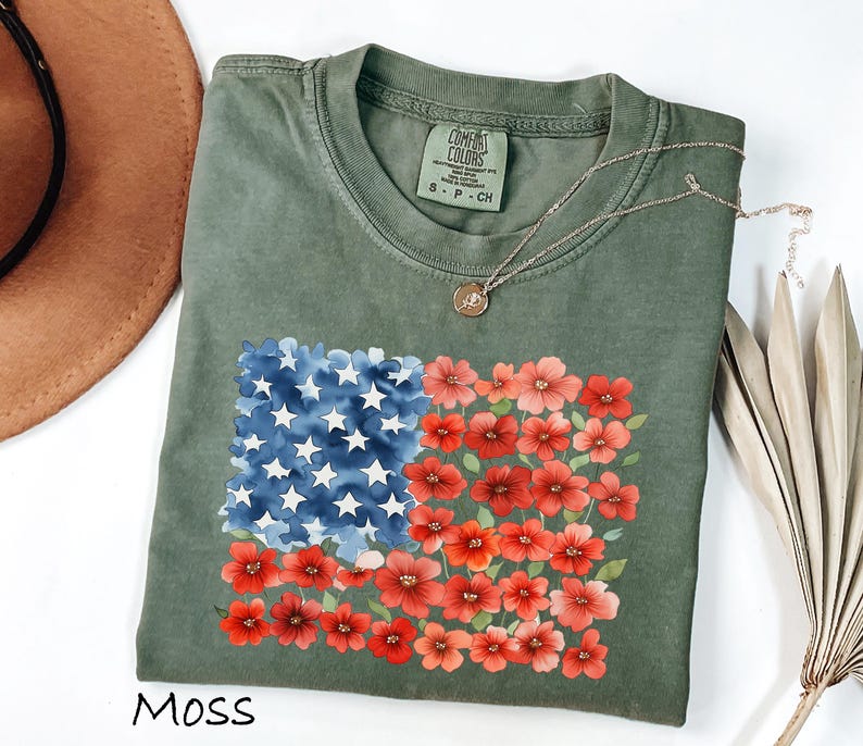 May include: Moss green t-shirt with a floral American flag design. The flag design features blue watercolor stars and red floral stripes. The shirt has a crew neck and short sleeves. A gold necklace is draped across the shirt.