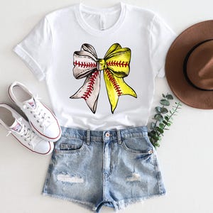 May include: White t-shirt with a yellow and white softball bow graphic. The bow is tied with a red ribbon. The shirt is paired with denim shorts and white sneakers.