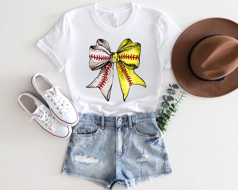 May include: White t-shirt with a yellow and white softball bow graphic. The bow is tied with a red ribbon. The shirt is paired with denim shorts and white sneakers.