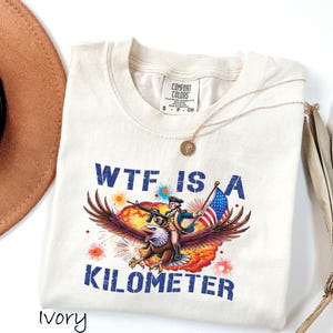 K&ouml;nnte beinhalten: Cremefarbenes T-Shirt mit einem Aufdruck eines Adlers, der einen Soldaten mit Gewehr und amerikanischer Flagge tr&auml;gt. Der Text "WTF IS A KILOMETER" ist blau gedruckt. Eine goldene Halskette und ein brauner Hut sind ebenfalls zu sehen.