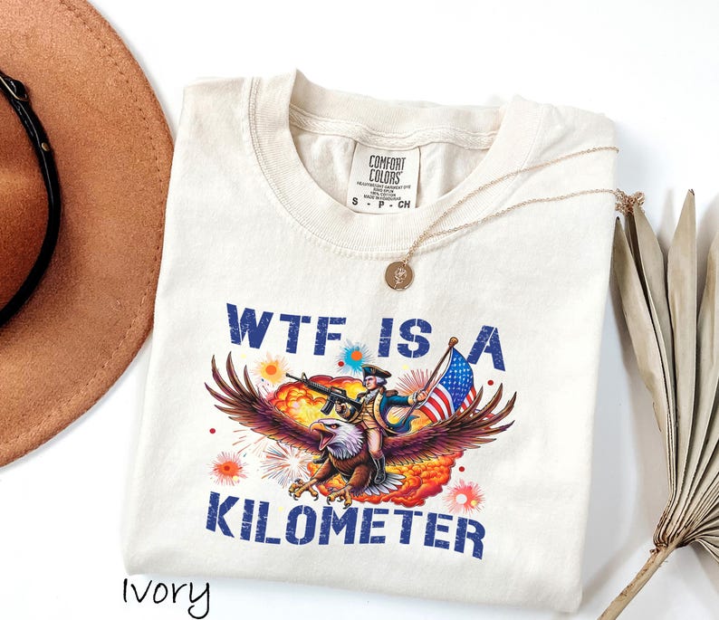K&ouml;nnte beinhalten: Cremefarbenes T-Shirt mit einem Aufdruck eines Adlers, der einen Soldaten mit Gewehr und amerikanischer Flagge tr&auml;gt. Der Text "WTF IS A KILOMETER" ist blau gedruckt. Eine goldene Halskette und ein brauner Hut sind ebenfalls zu sehen.