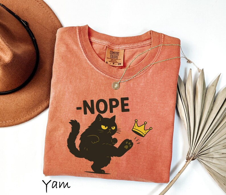 May include: A coral-colored t-shirt with the word "-NOPE" above a black cat kicking a gold crown. The cat has yellow eyes and a grumpy expression. A gold necklace and a brown hat are also in the image.