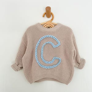 May include: A beige knitted jumper with a blue embroidered letter "C" on the front.