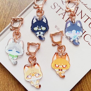 May include: Five acrylic keychains featuring stylized cat illustrations in various colors including dark blue, light gray, orange, and tan. Each charm is attached to a rose gold-colored metal cat-shaped clasp and ring hardware.