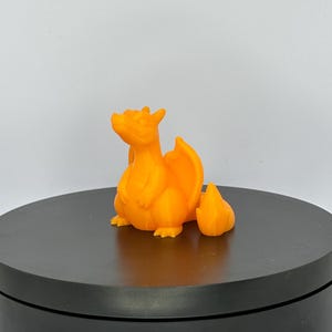 Può includere: Una statuetta di drago arancione brillante, stampata in 3D, con una fiamma. Il drago ha grandi ali ed è seduto in posizione eretta. La statuetta è su un espositore circolare nero.
