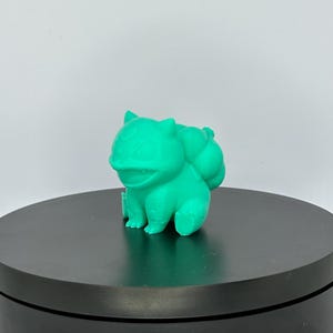 Può includere: Una statuetta di Snorlax, un personaggio di Pokemon, stampata in 3D in verde turchese, seduta su un giradischi nero.