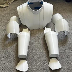 May include: A set of white 3D printed armor components including a central chest piece with a circular neck opening, two shoulder guards, two upper arm pieces, two forearm guards, and two hand plates, all featuring a smooth, rigid plastic finish.
