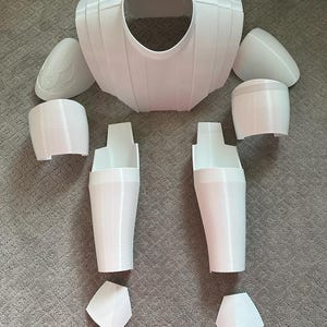May include: A disassembled white plastic costume, including a chest piece, shoulder pads, arm guards, leg guards, and knee pads. The pieces are arranged on a gray surface, ready for assembly. The costume appears to be 3D printed.