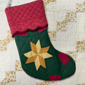 May include: A quilted Christmas stocking with a green background and a large yellow star in the center. The stocking is trimmed with red fabric and has a scalloped edge.