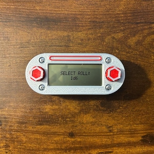 May include: A rectangular, silver-colored electronic device with a small screen displaying "SELECT ROLL: 1d6." The device has red hexagonal buttons on each end and is secured with screws. A red stripe accents the top.