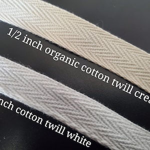 May include: Two samples of 1/2 inch cotton twill ribbon. The top ribbon is cream colored and labeled "1/2 inch organic cotton twill cream". The bottom ribbon is white and labeled "1/2 inch cotton twill white".