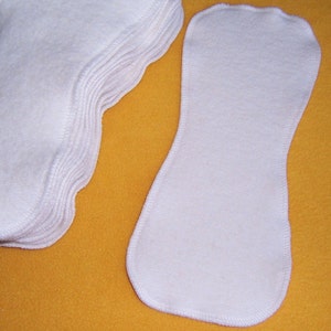 May include: A stack of white cloth diaper inserts, with one insert laid out flat on a yellow surface. The inserts are made of a soft, absorbent material and are designed to be used with cloth diapers.