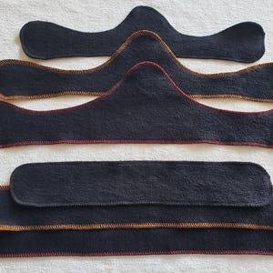May include: A collection of dark navy blue fabric pieces with contrasting stitching. The pieces vary in shape, including curved and rectangular designs, with red and gold stitching outlining the edges. The items are arranged on a white surface.