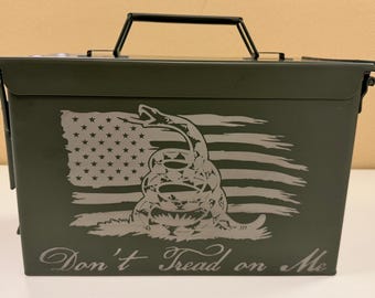 Pode incluir: Uma caixa de munição de metal verde-oliva com uma alça preta. A frente apresenta um design de bandeira americana desgastada com uma cobra enrolada e as palavras "Don't Tread on Me" em uma fonte de script. A caixa tem uma tampa articulada e um fecho de trava.