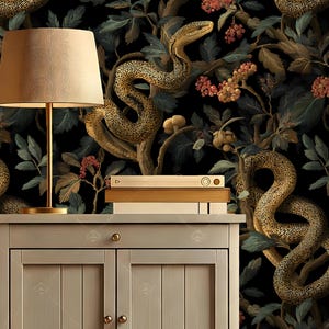 May include: A bedroom scene featuring a nightstand with a lamp and books. The nightstand is light gray with two doors and gold hardware. The wallpaper behind the nightstand has a dark background with a pattern of snakes, leaves, and berries.