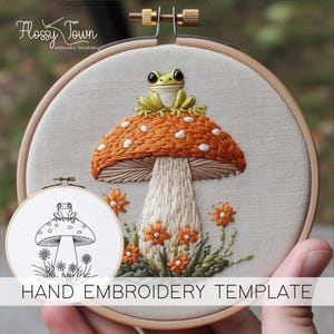 May include: Hand embroidery template featuring a green frog sitting on a mushroom with orange and white accents. The mushroom is surrounded by green leaves and orange flowers. The template is on a white fabric background.