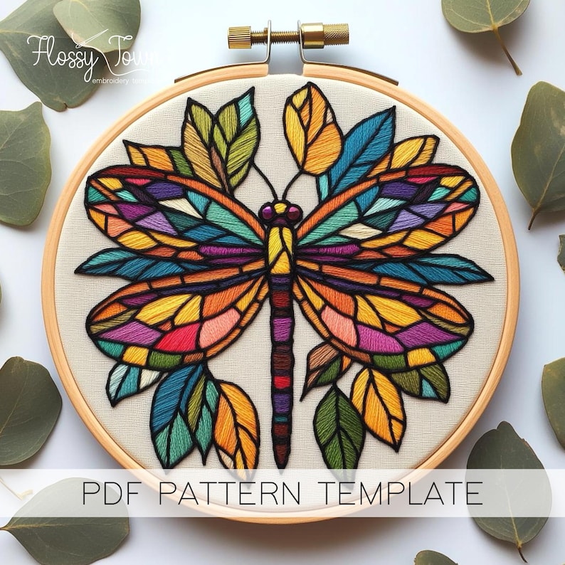 May include: A colourful stained glass style dragonfly embroidery pattern in a wooden embroidery hoop. The dragonfly is surrounded by leaves in shades of green, yellow, and blue.
