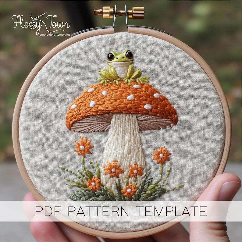 May include: Embroidery pattern template featuring a green frog sitting on top of an orange and white mushroom with white dots. The mushroom is surrounded by green leaves and orange flowers. The pattern is displayed in a wooden embroidery hoop.