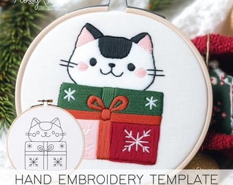 Könnte beinhalten: Handstickvorlage mit einer weißen Katze mit schwarzem Fleck, die aus einer grünen und roten Geschenkbox kommt. Das Design beinhaltet Schneeflocken und eine Schleife. Der Text "HAND EMBROIDERY TEMPLATE" befindet sich unten.
