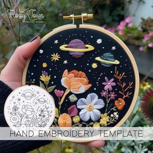 May include: A black embroidery hoop with a hand-stitched design of flowers and planets on a dark blue background. The design includes a large orange flower, a white flower, and two planets with rings. The text "HAND EMBROIDERY TEMPLATE" is printed below the hoop.
