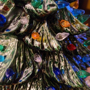 May include: A ceramic Christmas tree with a green glaze and a textured bark-like surface. The tree is decorated with colorful lights, including red, blue, green, yellow, and clear.