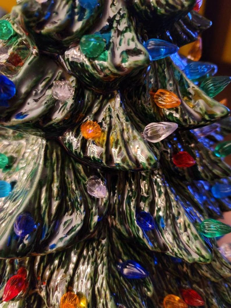 May include: A ceramic Christmas tree with a green glaze and a textured bark-like surface. The tree is decorated with colorful lights, including red, blue, green, yellow, and clear.