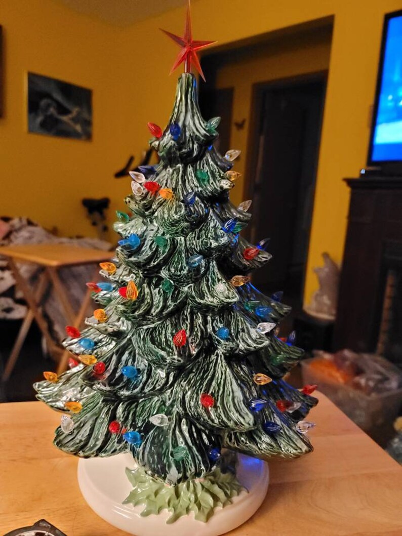 May include: A ceramic Christmas tree with a green glaze and a white base. The tree is decorated with colorful lights and a red star on top.