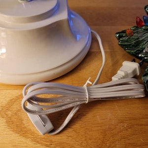 May include: A white ceramic base with a black cord and a white plug. The cord has a switch on it. The base is for a small, artificial Christmas tree.
