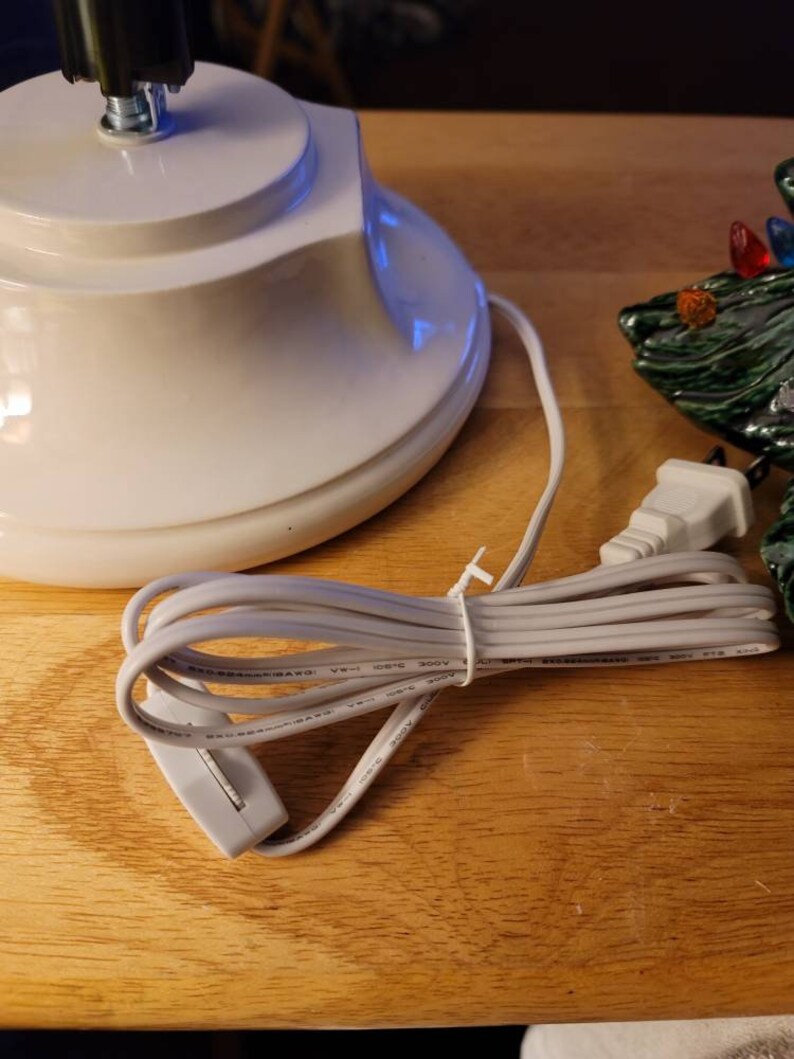 May include: A white ceramic base with a black cord and a white plug. The cord has a switch on it. The base is for a small, artificial Christmas tree.