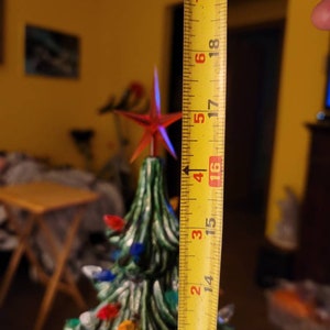 May include: A yellow measuring tape with red numbers is being used to measure a green ceramic Christmas tree with colorful lights. The tape measures 19 inches.
