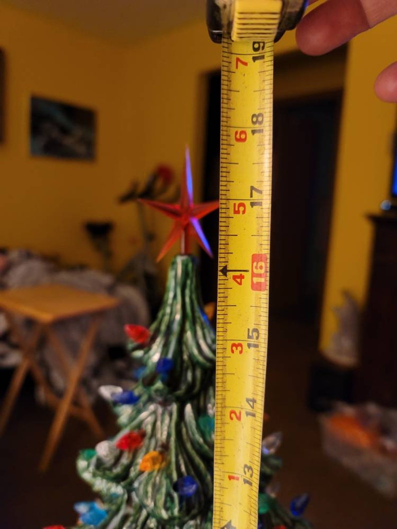 May include: A yellow measuring tape with red numbers is being used to measure a green ceramic Christmas tree with colorful lights. The tape measures 19 inches.
