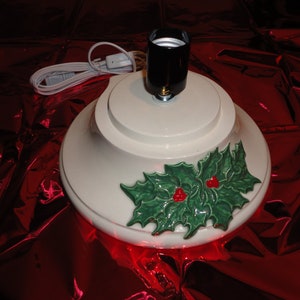 May include: White ceramic Christmas tree stand with a green holly leaf design and red berries. The stand has a black light socket and a white cord.